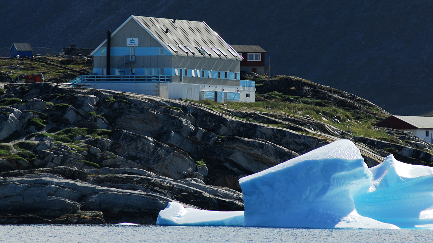 Hotel Disko Bay på klippekant med isbjerg i forgrunden og bjerg i baggrunden.