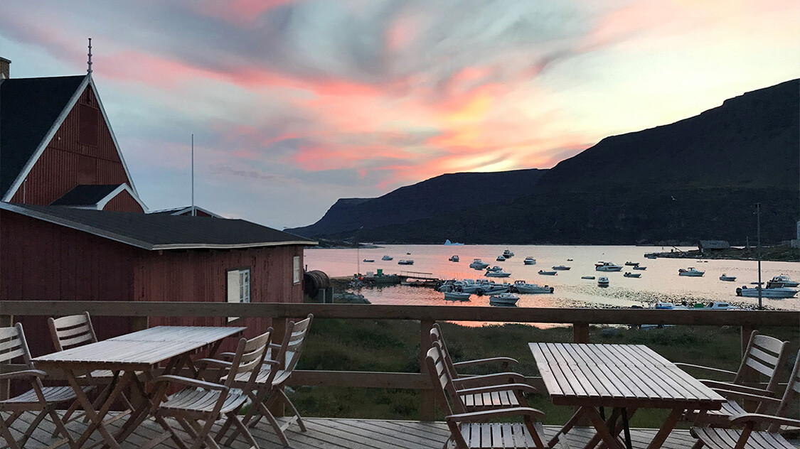 Solopgang på terrasse på Hotel Disko Island på Diskoøen med udsigt over havn med både. 