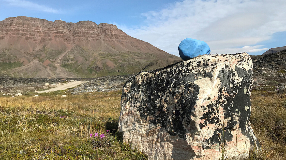 Pejlemærke til vandrere, bestående af blåmalet sten placeret ovenpå større klippe. Ved siden af klippen ses nogle små blomster, og i baggrunden et massivt fjeld med et fjernt tårn på toppen. 