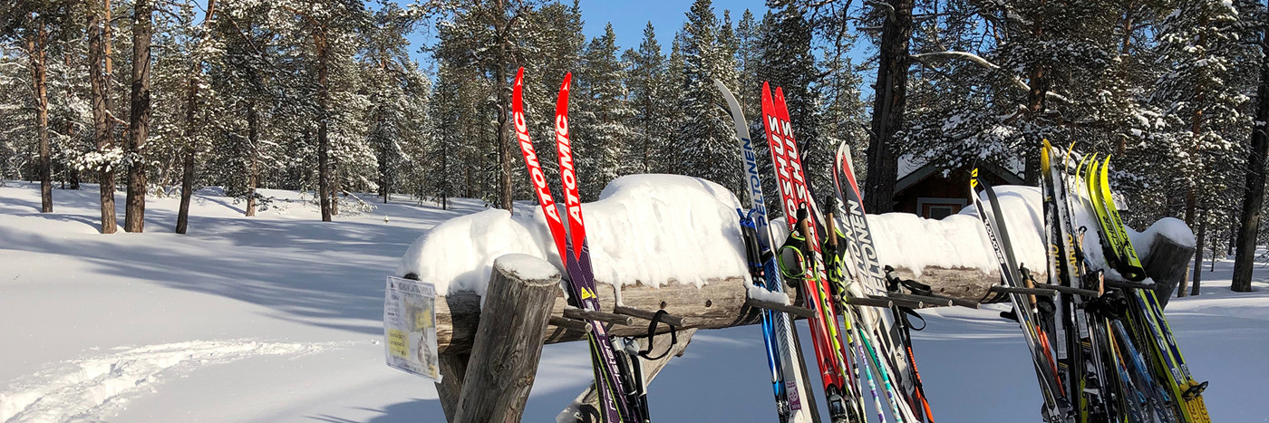 Adskillige par af ski, stillet op mod en vandret pæl med et tykt lag sne oven på. I baggrunden ses en sneklædt nåleskov og hytte. 