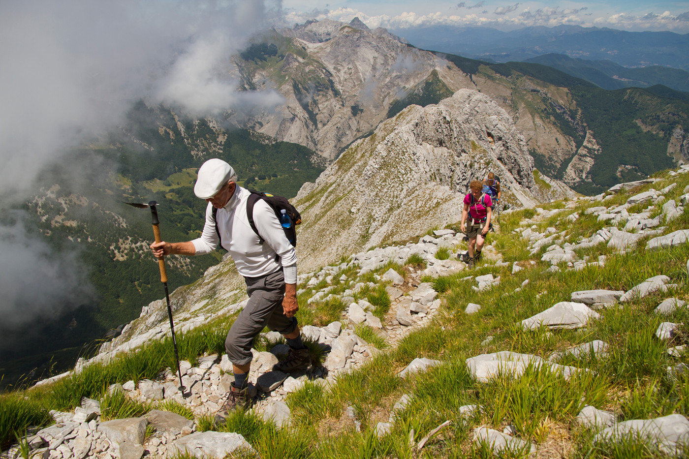 3 personer med rygsække og vandrestave vandrer langs stejl bjergryg med udsigt over de Toscanske Appenniner, med hvide skyer på himlen og frodige, grønne skove i dalen nedenfor.  