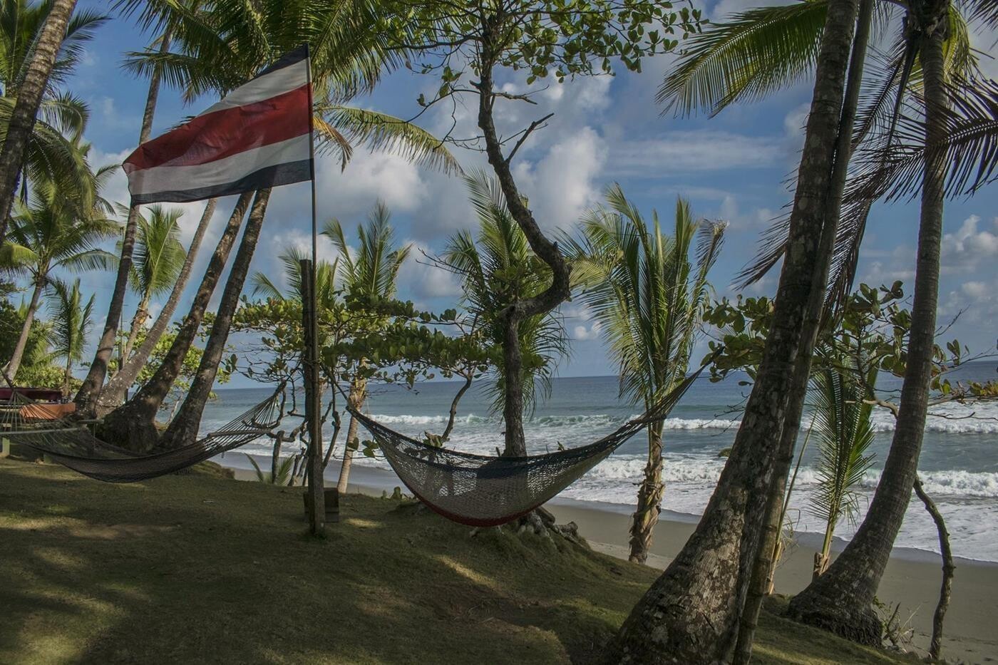 Hængekøjer ophængt fra palmetræer på sandstrand med brusende bølger i baggrunden. Costa Ricas flag blæser i vinden. 