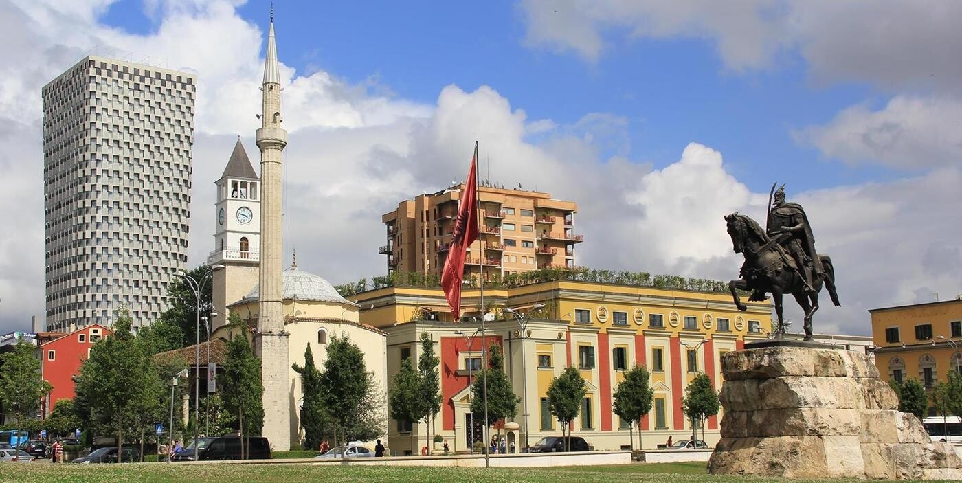 Statue af rytter på hest i forgrunden, med albanske bygninger i baggrunden.