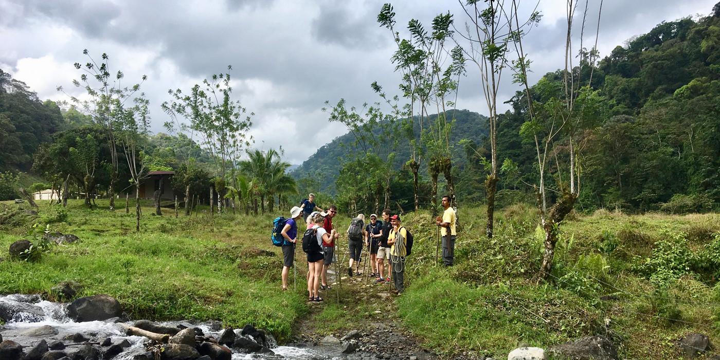 Gruppe af vandrere med guides står ved kanten til en sprudlende flod, med regnskov og bjerge i baggrunden.