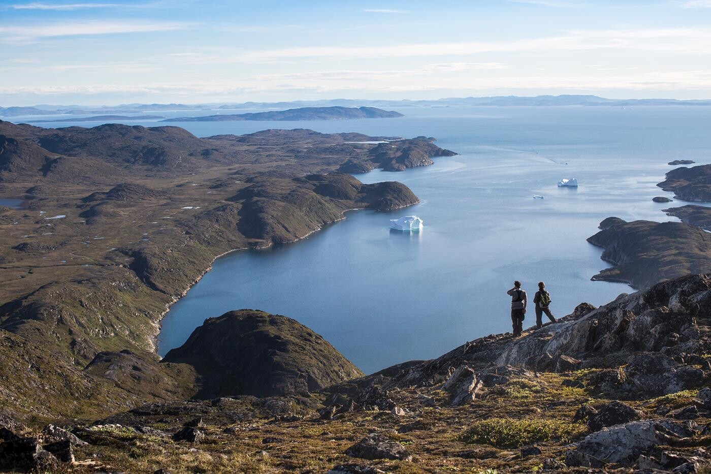 To personer på vandretur i forgrunden, som beundrer udsigten over Diskobugten i Grønland, med små isbjerge i fjorden nedenfor. 