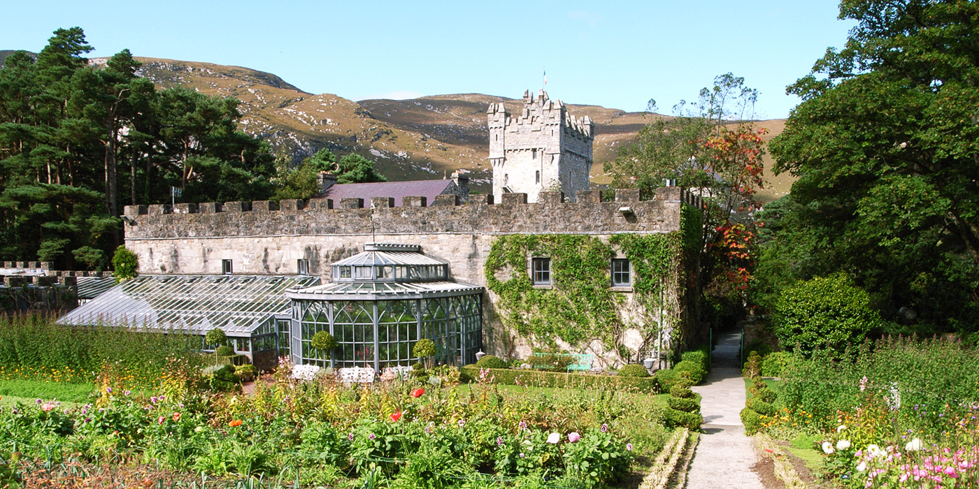 Glenveagh Castle; et slot af sten med brystværn langs muren, et borg-lignende tårn, og drivhuse. I forgrunden ses et blomsterbed, og i baggrunden ses irske, golde bjerge.