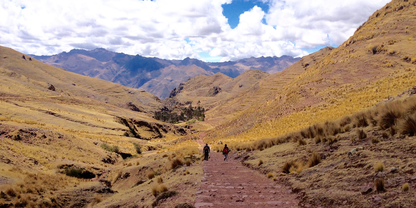To personer trekker gennem Den Hellige Dal, med rødlig vegetation langs bjergsiderne på begge sider. Et stort bjergmassiv strækker sig langs hele horisonten. 