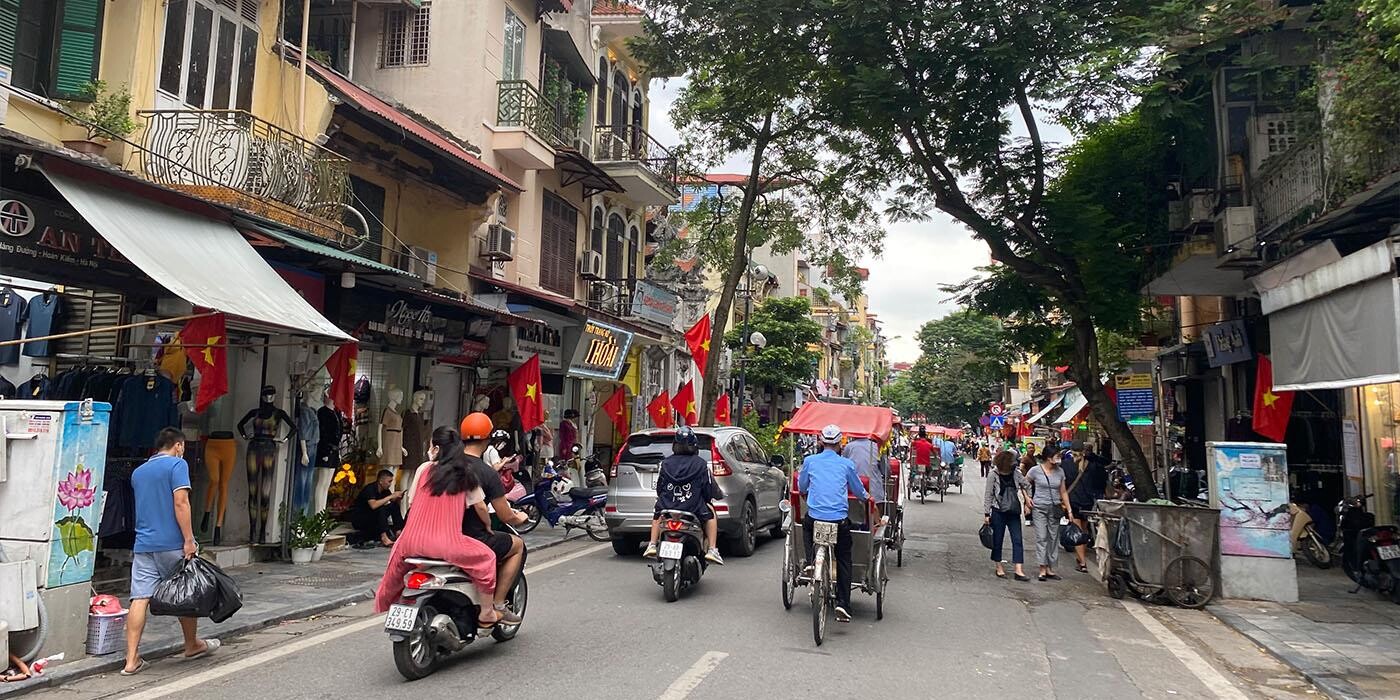 Travl gade i Hanoi med butikker, fodgængere, scootere, cykler og biler. Der er mange vietnamesiske flag. 