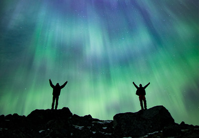 Silhuet af to personer på klippetop, med armene strakte i vejret, mod himmel fuld af stjerner og grønt og blåt nordlys. 