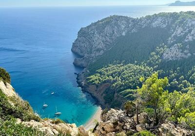 Bugt og sandstrand på Mallorca med 3 både i det turkisblå hav, omgivet af bjerge dækket af træer og buske