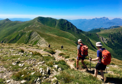 To personer med vandreudstyr i forgrunden, med flere vandrere længere væk og udsigt over Appeninnerne, med Apuanebjergene i horisonten. 