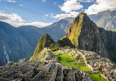 Udsigt over Machu Picchu. Velbevaret ruin på bjergtop med mange husmure i forskellige niveau er veltrimmede græsplæner med enkelte træeer. I baggrunden ses stor bjergryg. 