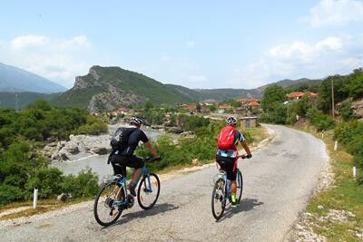 Cyklister kører ned af landevej med udsigt over flod og bjerge.