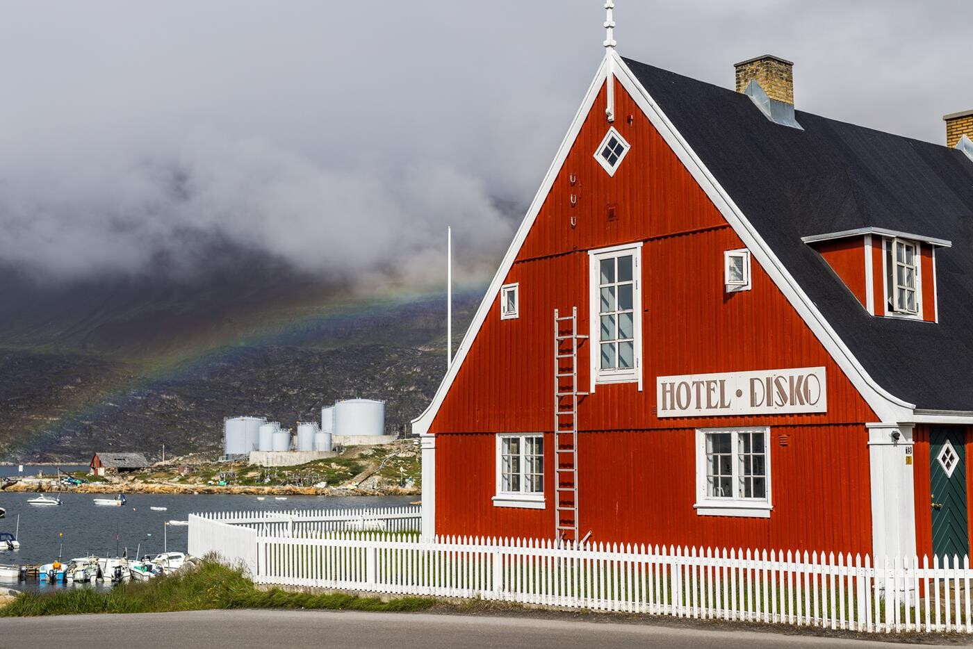 The main building of Hotel Disko Island, painted in bright red and white, with a view of the Disko Bay. A rainbow can be seen in the background.