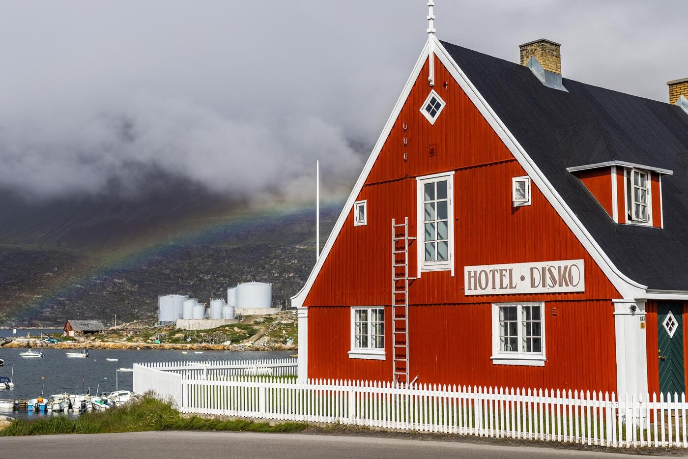 Hotel Disko Islands hovedbygning malet i rødt og hvidt, med udsigt over bugten. En regnbue ses i baggrunden. 